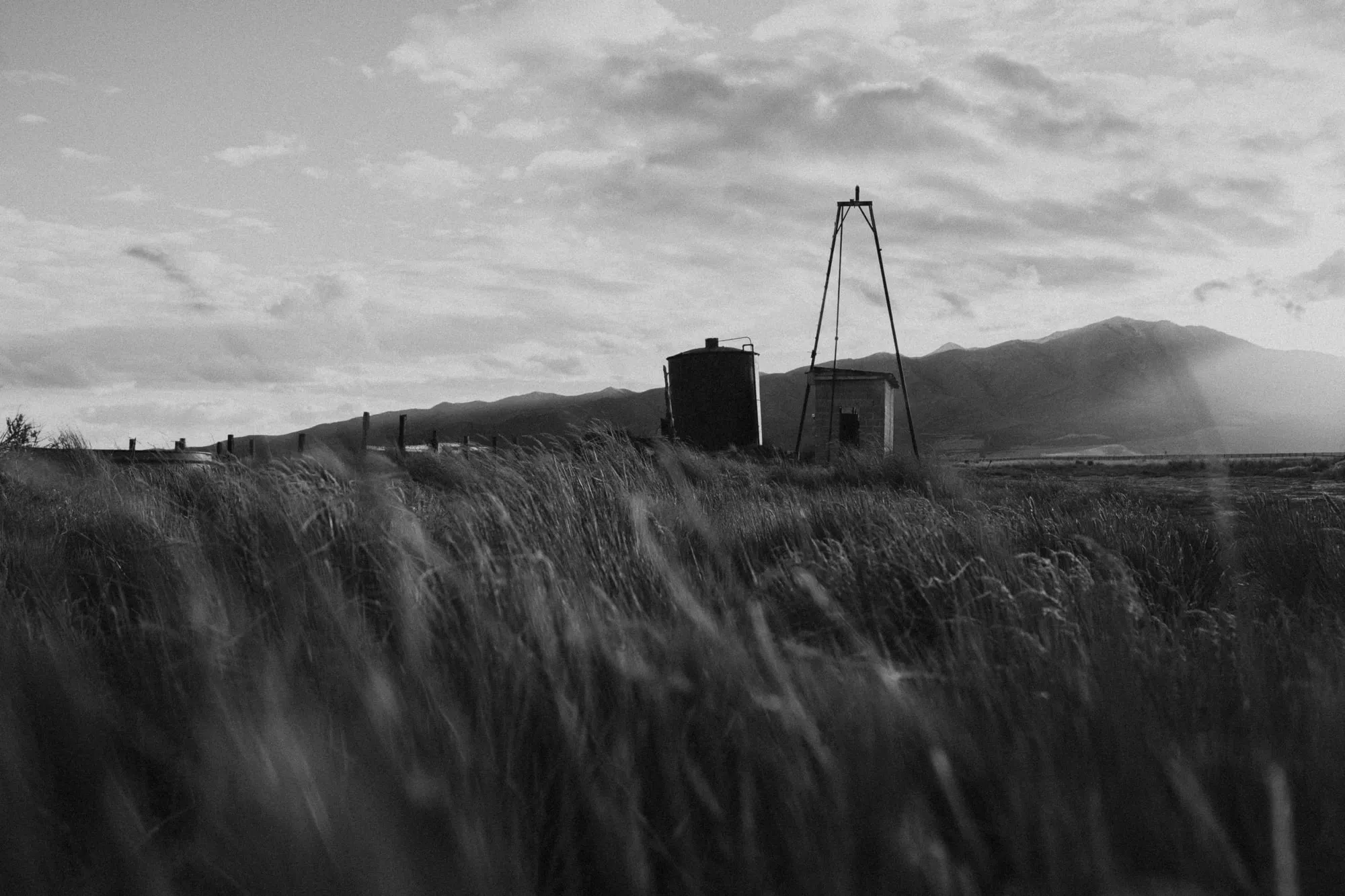 A steel water tank, a small outbuilding and a windmill derrick standing in wind-blown grass, with a mountain ridge behind them